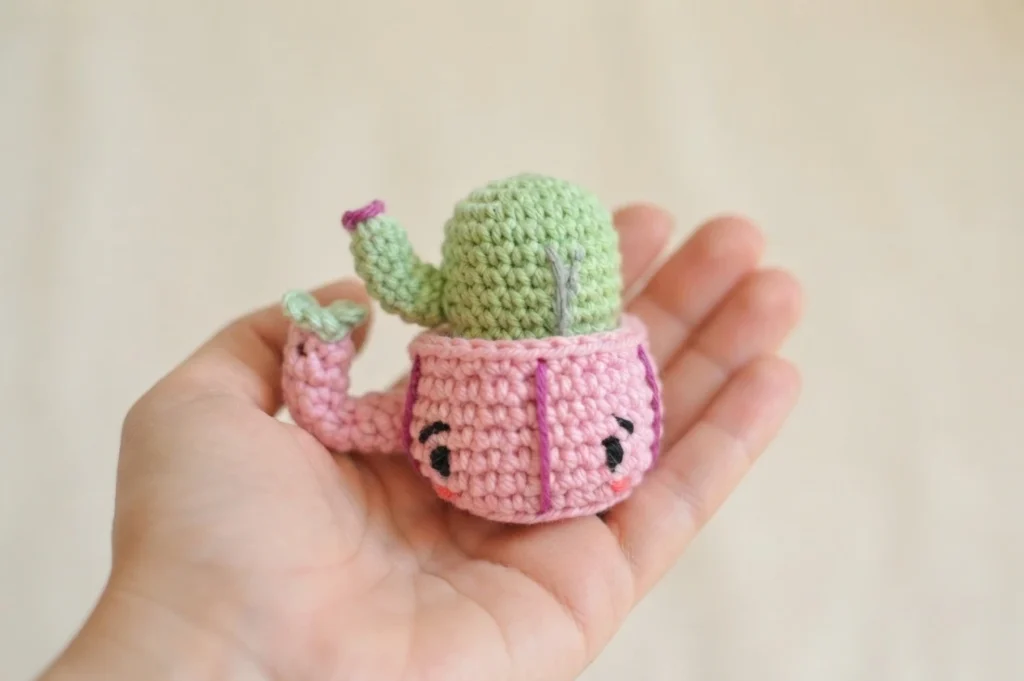 A hand holding a small pink crocheted mug with an embroidered smiling face, containing a green crocheted cactus with a tiny arm.
