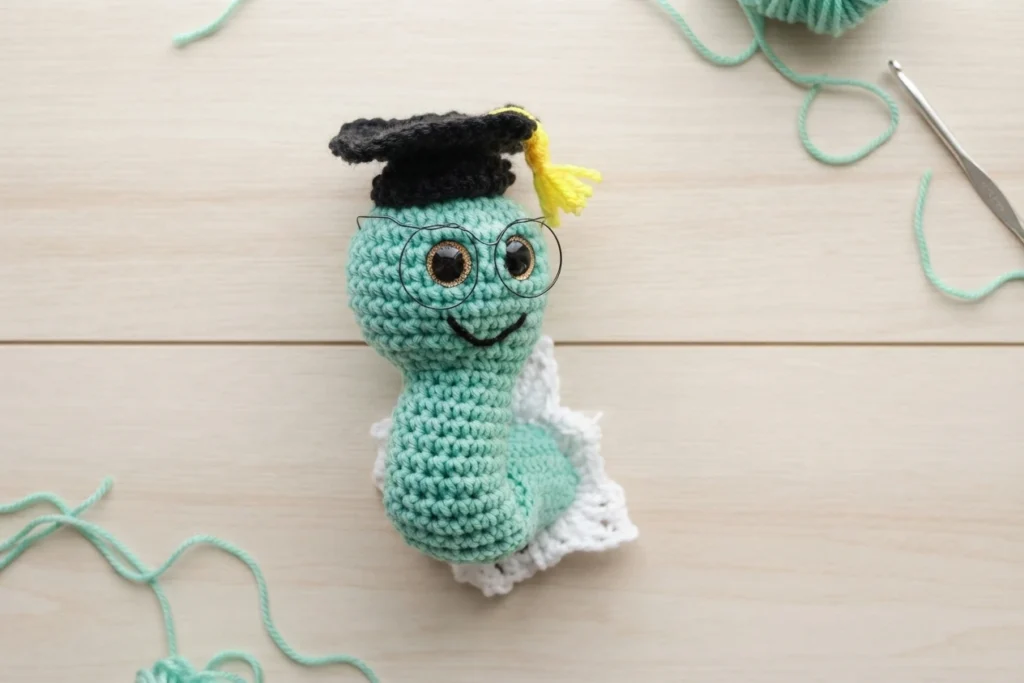 Crocheted mint green worm with a black graduation cap and yellow tassel, wire glasses and a stitched smile, resting on a white ruffled section on a light wood table with loose mint yarn and a crochet hook nearby.