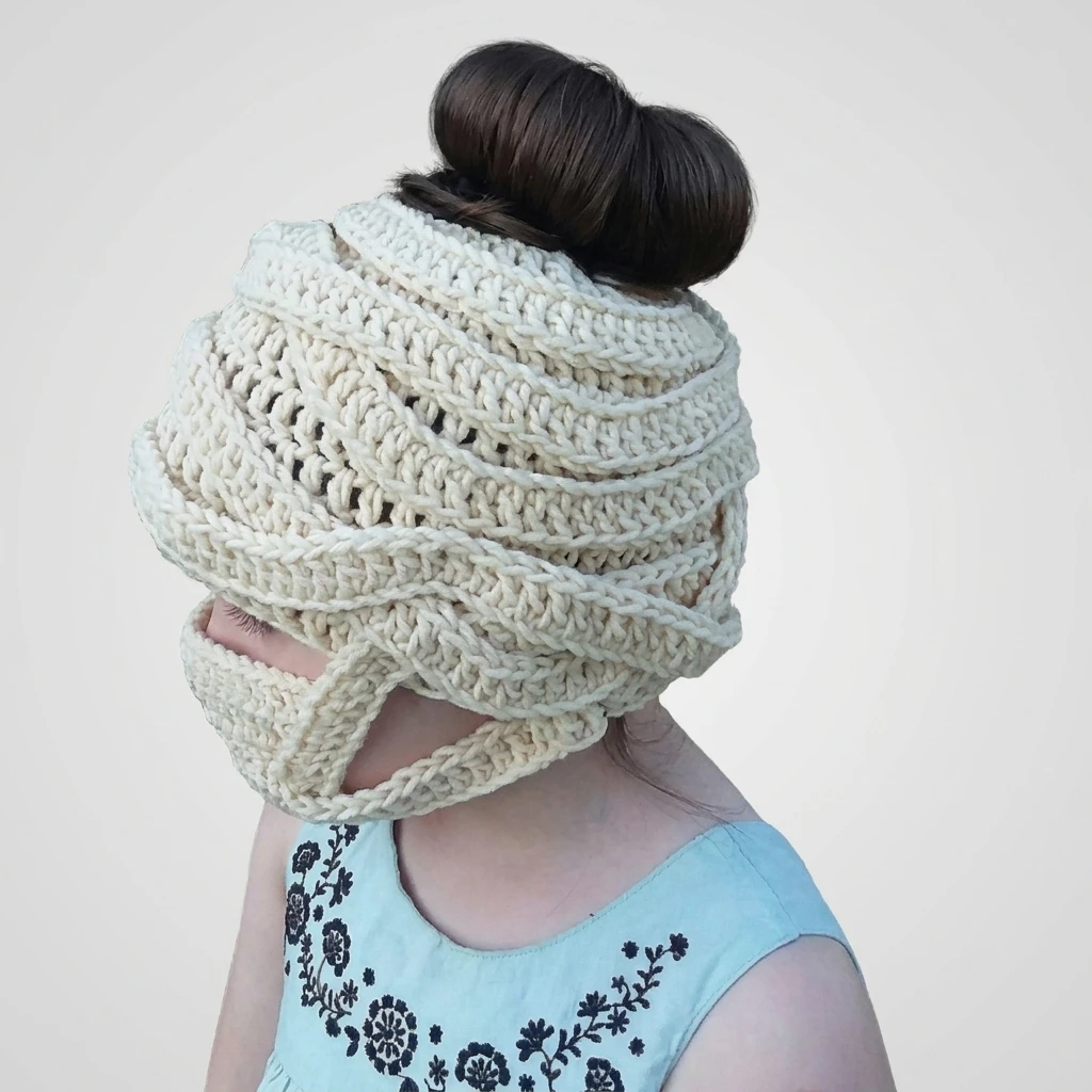 Child wearing a cream crochet mummy wrap beanie with the hair bun pulled through the top opening, wearing a light blue dress with dark floral embroidery.