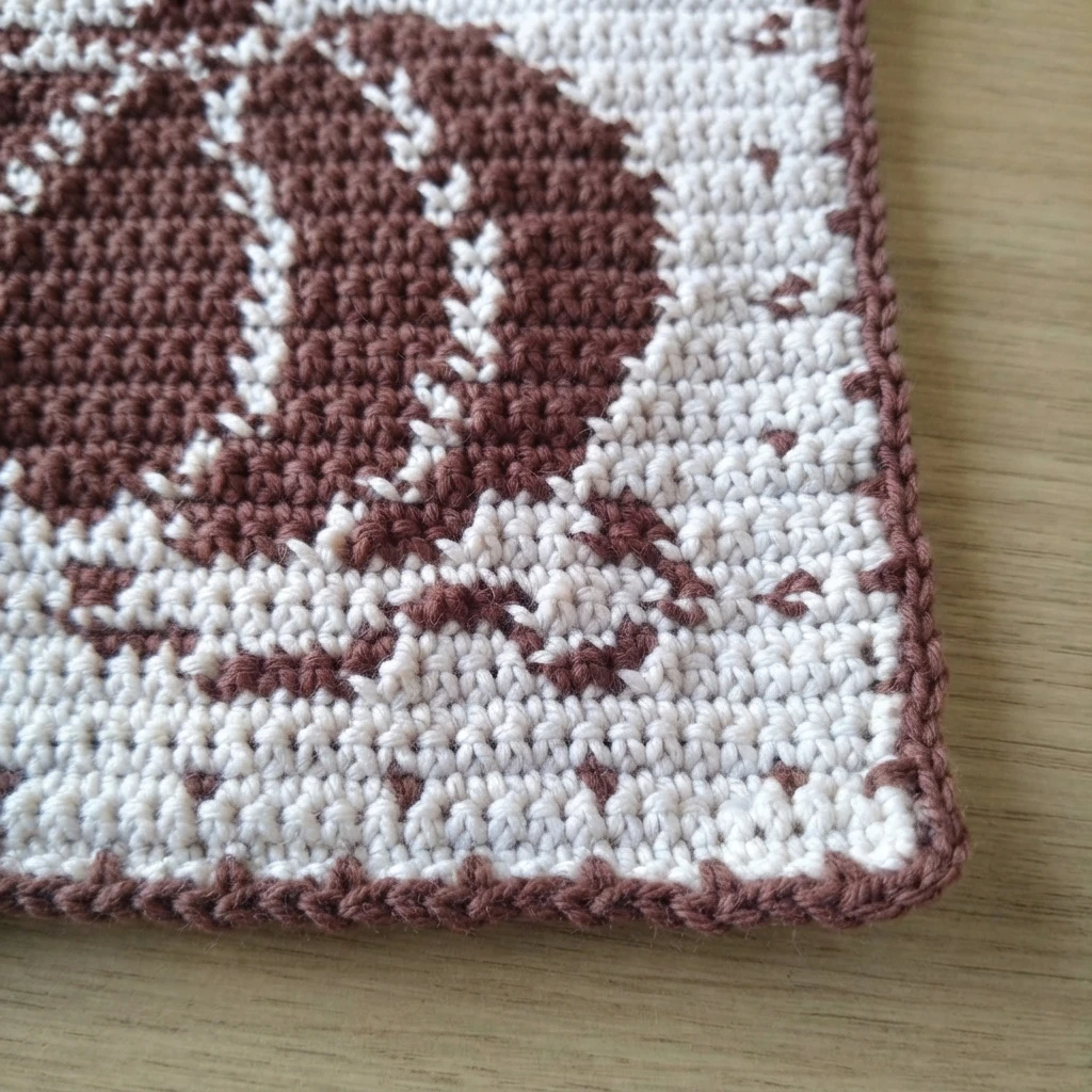 Gros plan du coin d’une manique crochetée crème et marron, avec une bordure marron autour d’un motif de citrouille.
