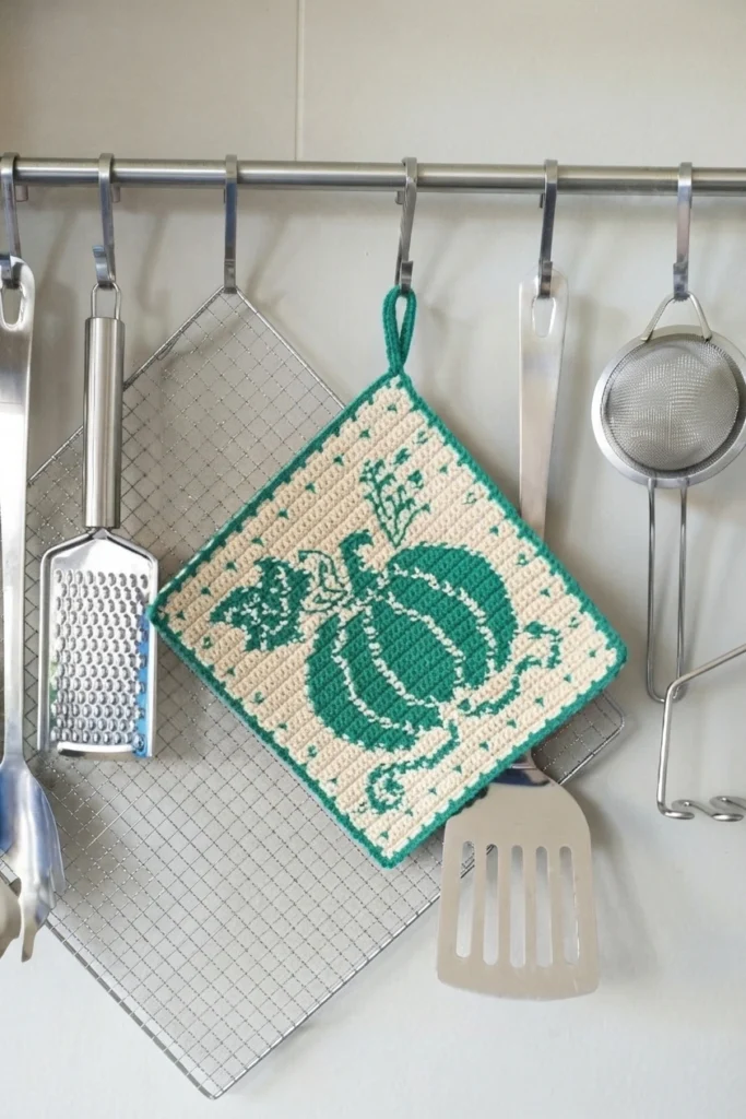 A cream and green crocheted pumpkin potholder hanging among kitchen utensils.