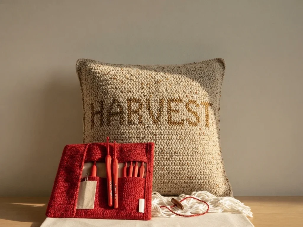 Finished cream tweed crochet pillow with the word harvest worked in tan letters, shown behind a red crocheted hook case holding red crochet hooks, with white yarn, a wooden button and a cream cloth on a wooden surface.