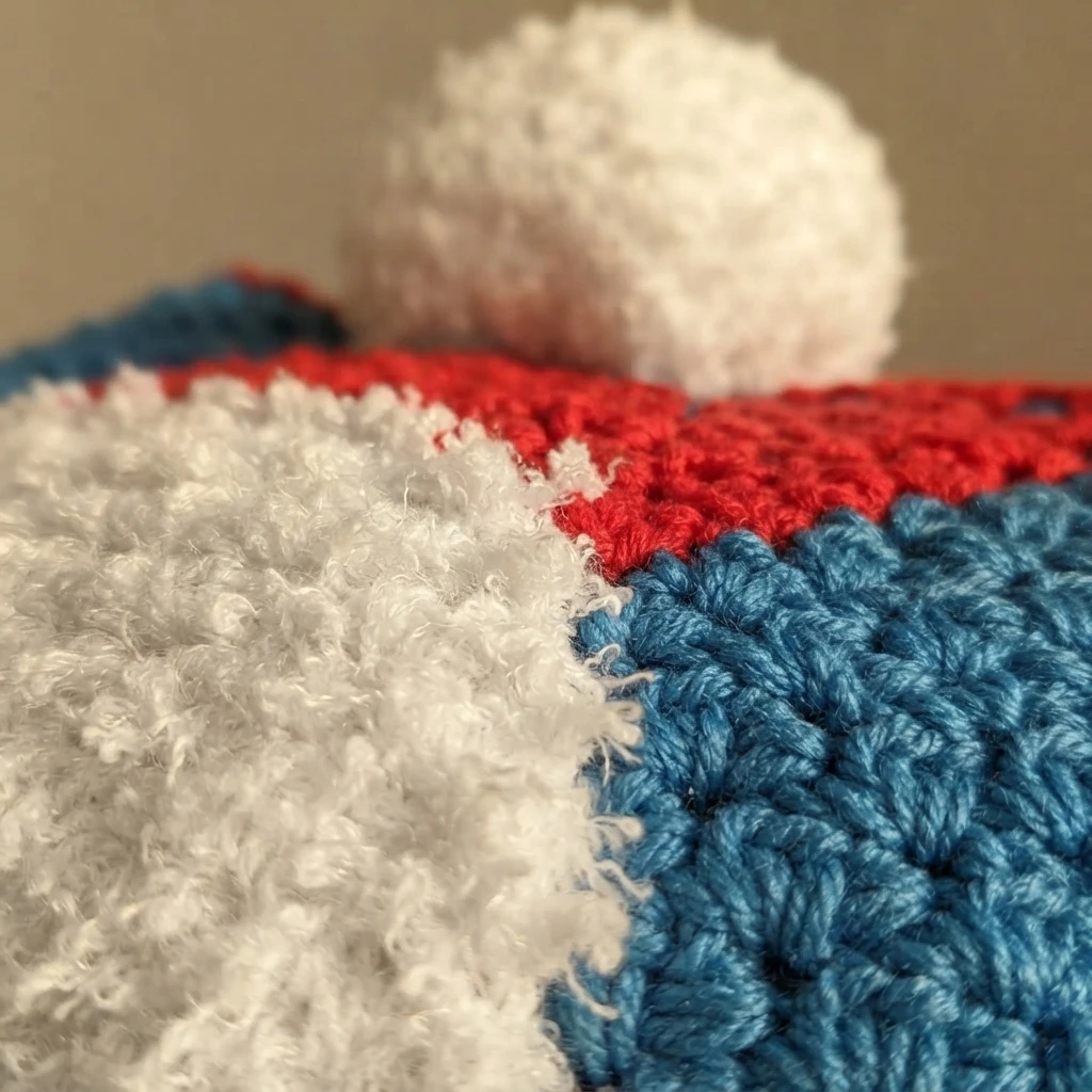 Close-up of a fuzzy white pom pom resting on red and blue corner to corner crochet stitches of a gnome pillow, with a white textured yarn section in the foreground.