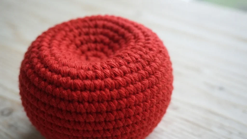 Red crocheted apple base with a deep, rounded indentation at the top, shown on a light wooden surface.