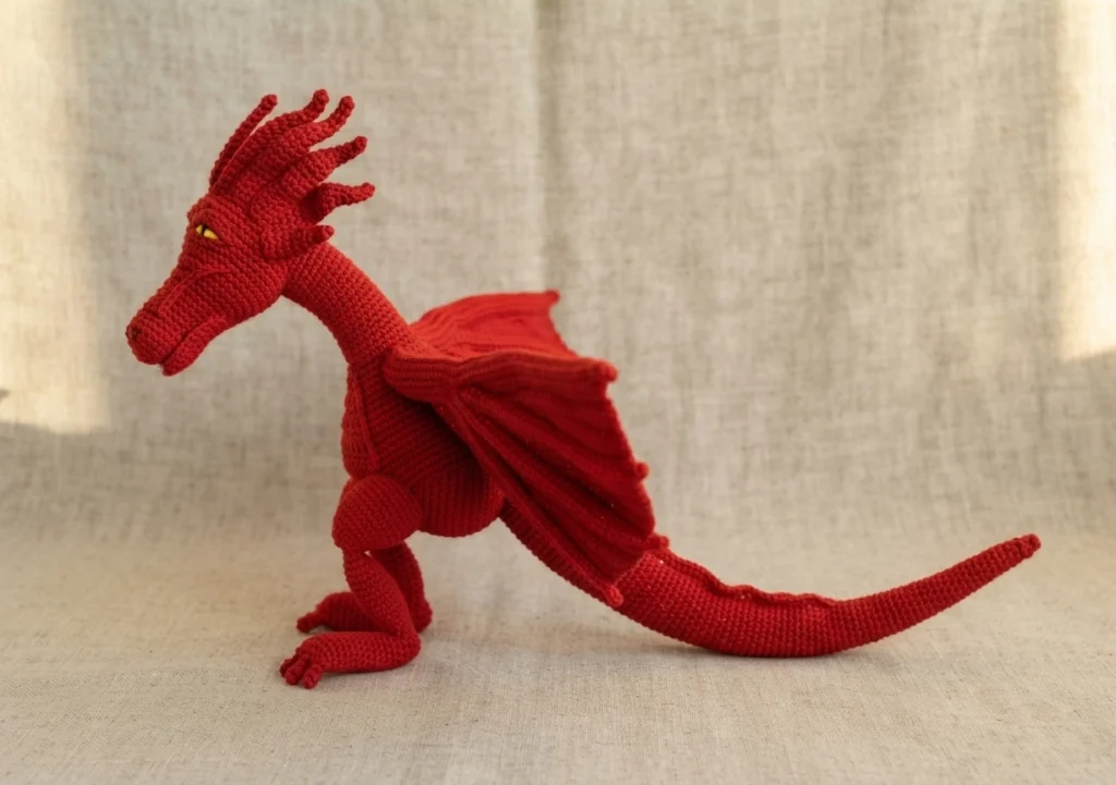 Red crochet dragon amigurumi shown in side profile with wings folded along its body and a long tail, on a neutral linen background.