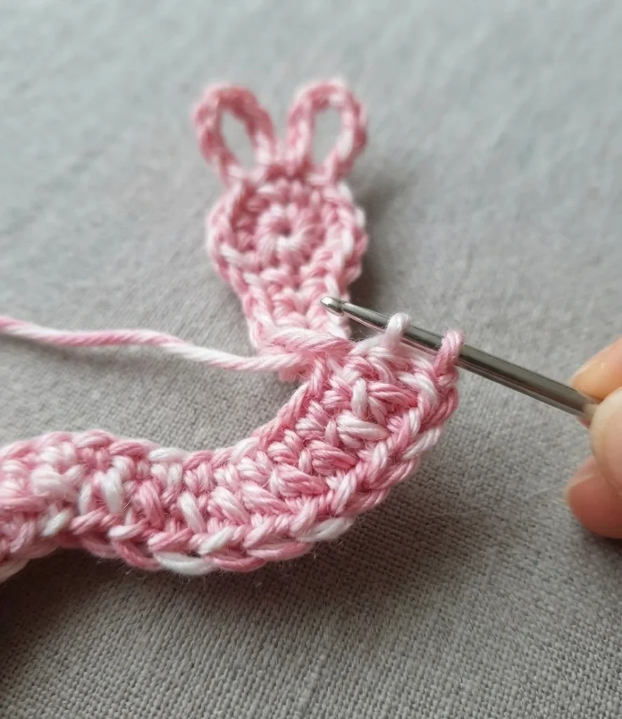 Pink and white crocheted bunny worry worm in progress on grey fabric, with a metal crochet hook inserted into the body and two looped ears at the top of the head.