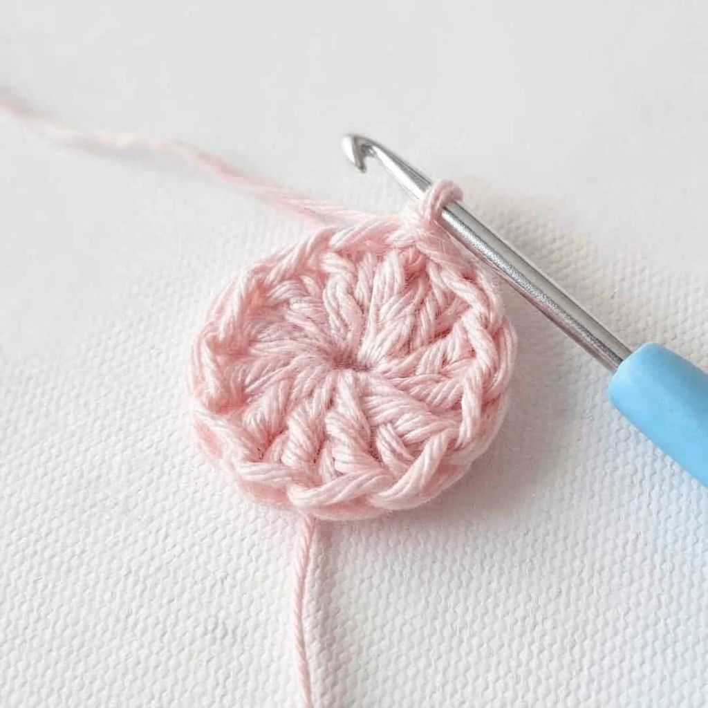 Pink crocheted circle in progress on a metal crochet hook with a light blue handle, resting on a white textured surface, with the working yarn looped on the hook and a long pink tail hanging down