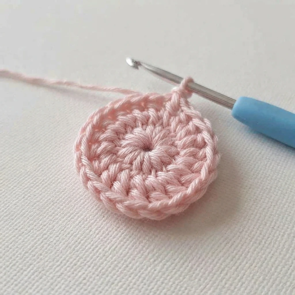 Pink crocheted circle of double crochet stitches on a metal hook with a blue handle, resting on light textured fabric, with the working yarn trailing to the left.