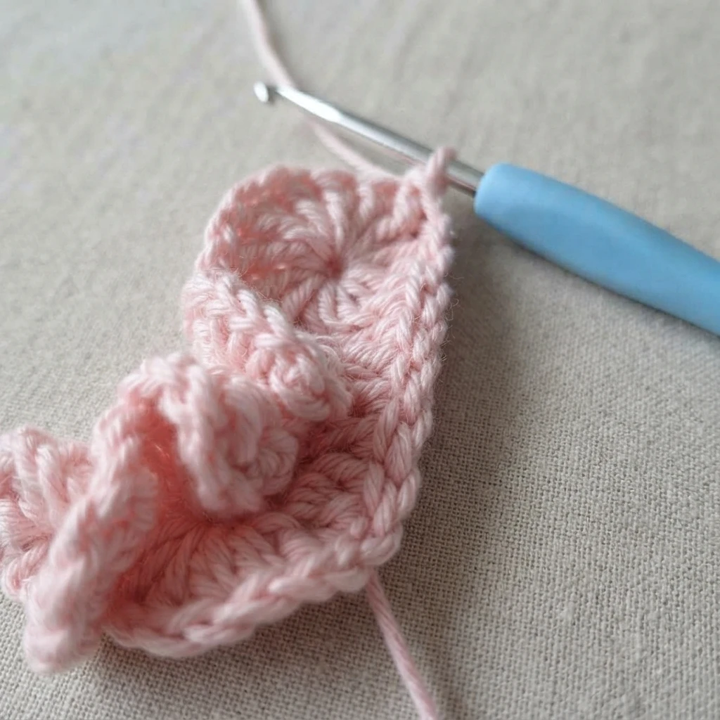 Pink crochet bunny head in progress on a blue handled crochet hook, resting on a light beige woven fabric with a loose yarn tail.