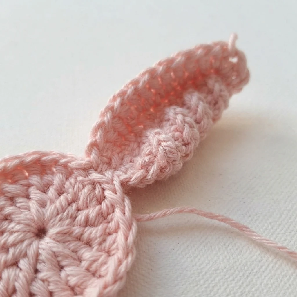 Close-up of a pink crochet piece in progress on a white surface, showing a round base worked in the round with a short textured strip extending from it, no hook visible.