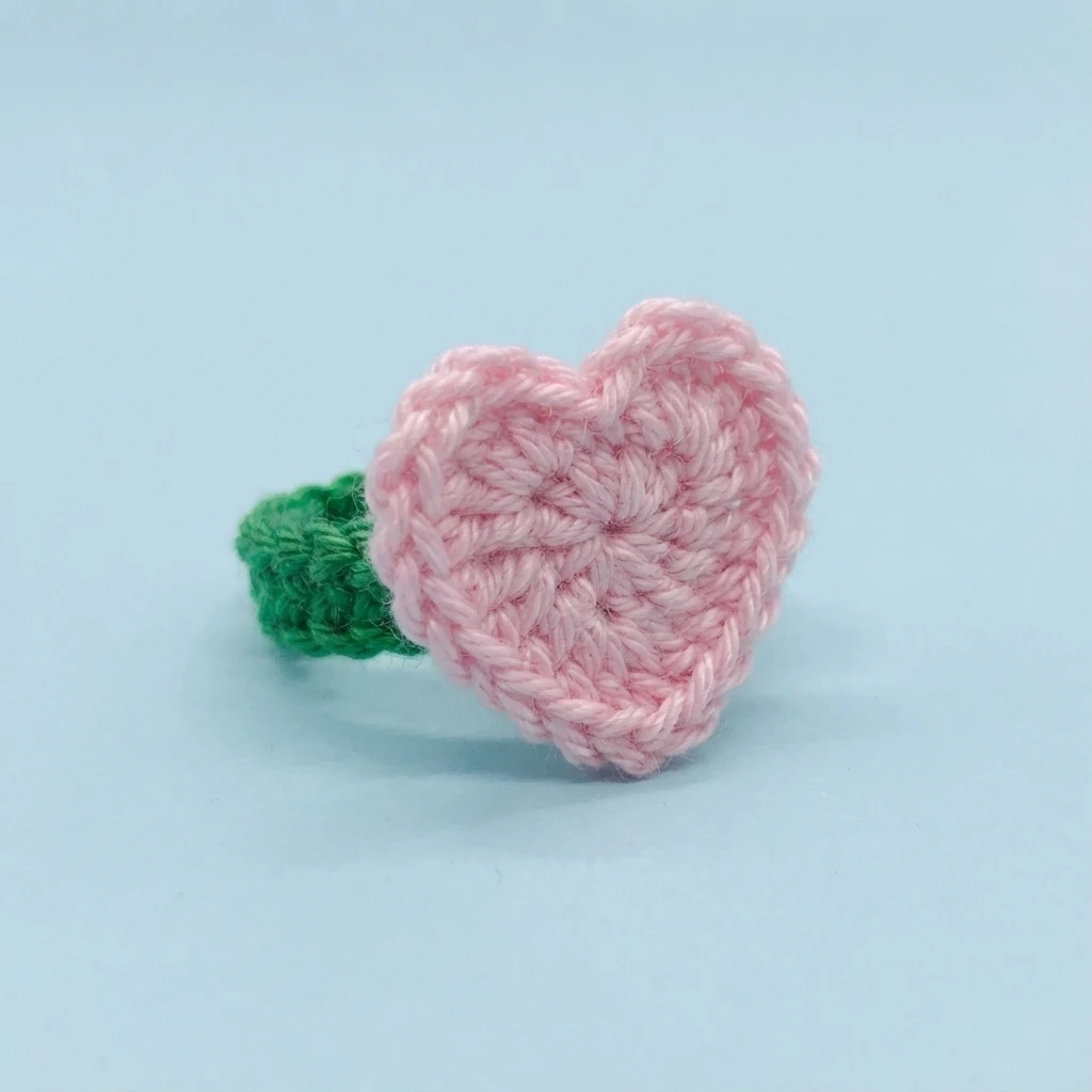 Crocheted ring with a light pink heart top and a green band on a light blue background