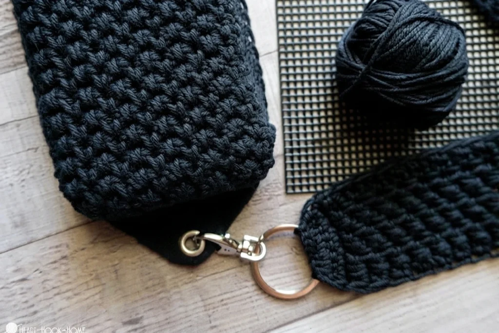 Close-up of a black crocheted bag and detached strap with metal clasp, beside black yarn and plastic canvas.