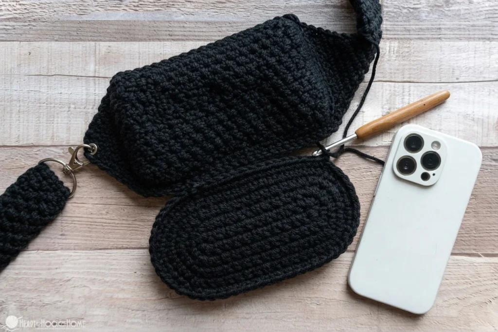 Black crocheted crossbody bag and strap laid out beside a white phone, with a crochet hook and loose yarn.