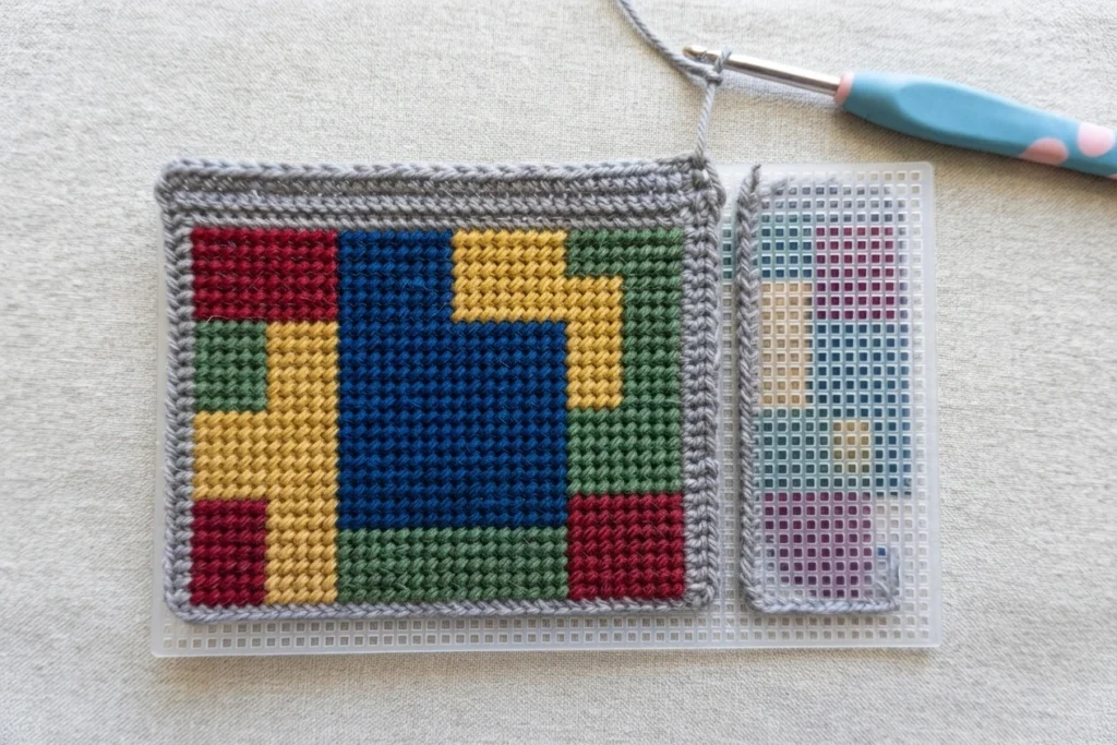 Panneau crocheté à motifs géométriques colorés posé sur une toile plastique, avec un crochet bleu à proximité.