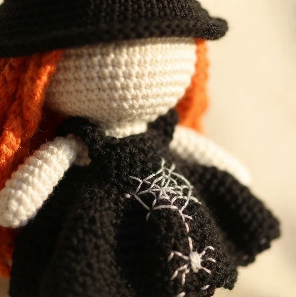 Close-up of a crocheted witch doll showing a black pointed hat, an ivory face, orange yarn hair and a black dress with white embroidered spiderweb and spider motifs.