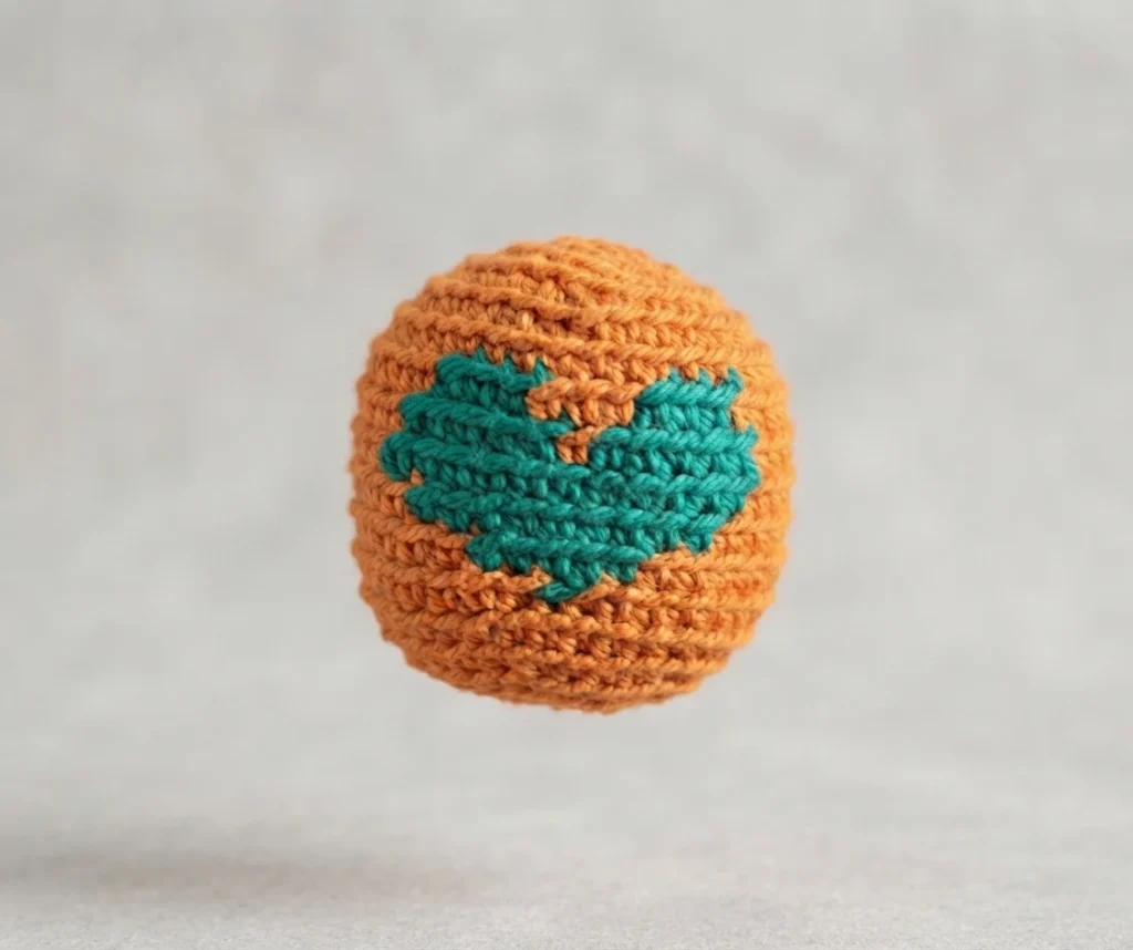 Single orange crocheted footbag with a teal heart on a light background
