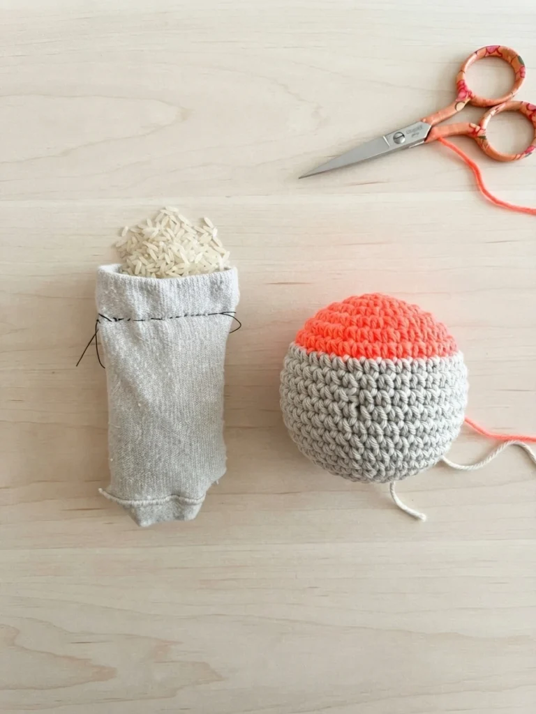 Sock filled with rice beside a two-tone grey and coral crocheted footbag ball with scissors on a light wooden surface.