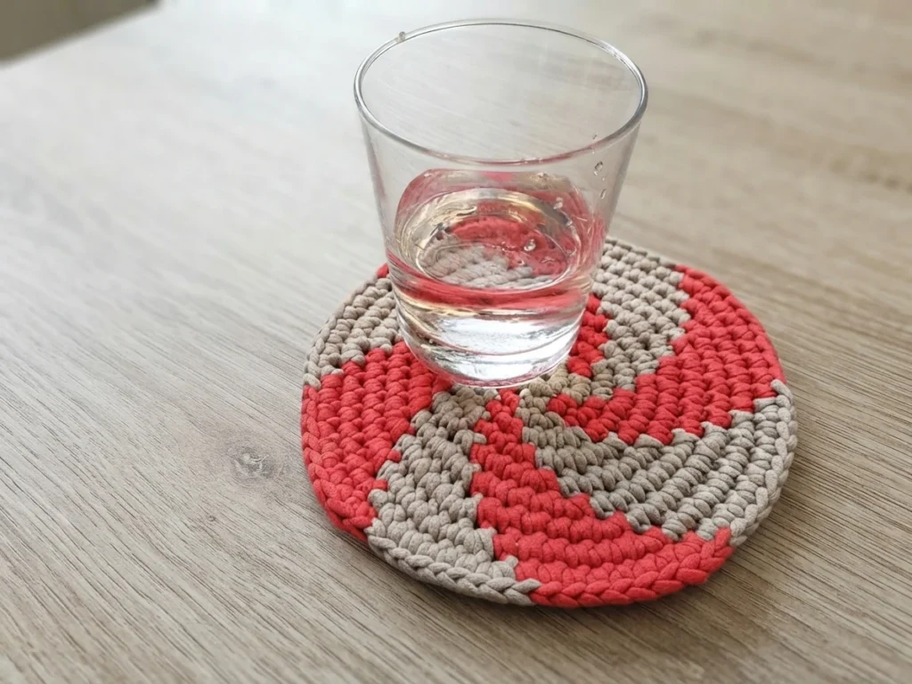 A glass of water rests on a round, chunky red-and-gray spiral crochet hot pad.