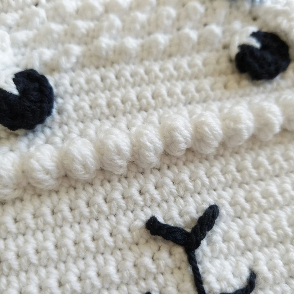 Close-up of a white crocheted lamb bib face showing the bumpy granule stitch trim, two round black and white eyes, and an embroidered black nose.