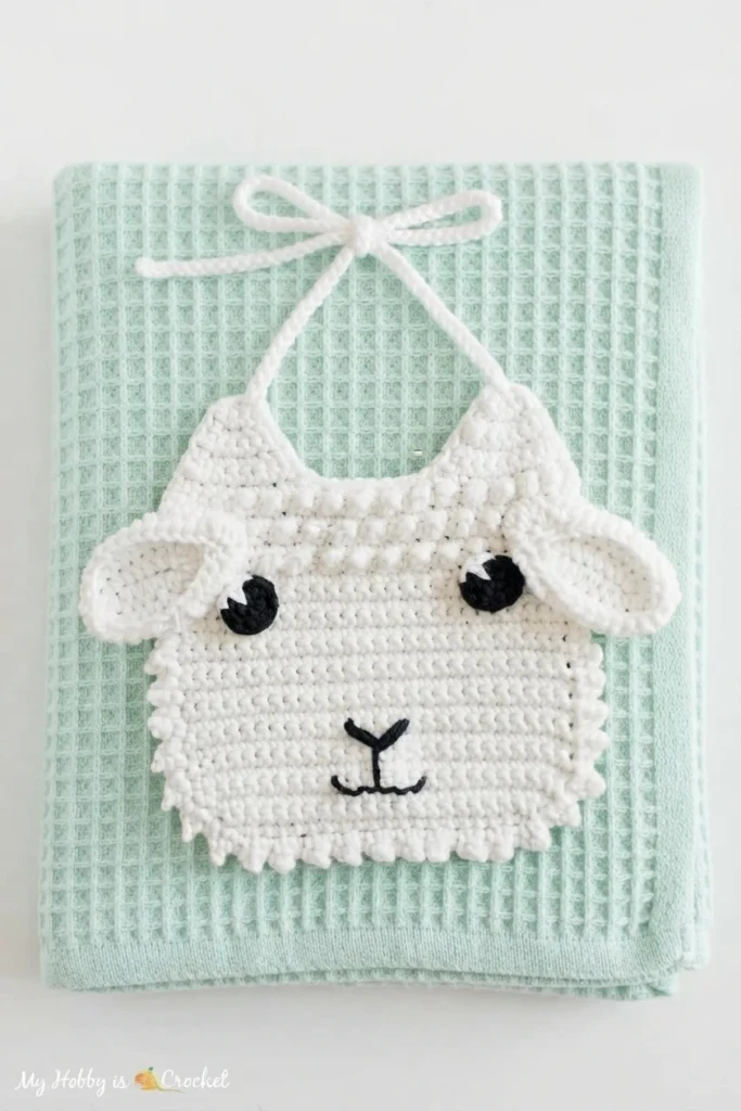 White crocheted lamb baby bib with black eyes, folded ears and embroidered nose, lying on a mint green waffle weave blanket.