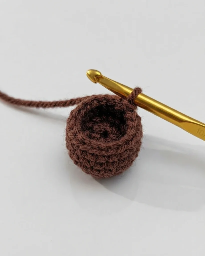 Petit ouvrage au crochet brun, ouvert et arrondi, tenu sur un crochet doré avec le fil attaché.
