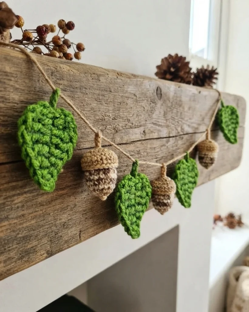 Crochet leaves and acorns hang from a beige cord against a rustic wooden board.