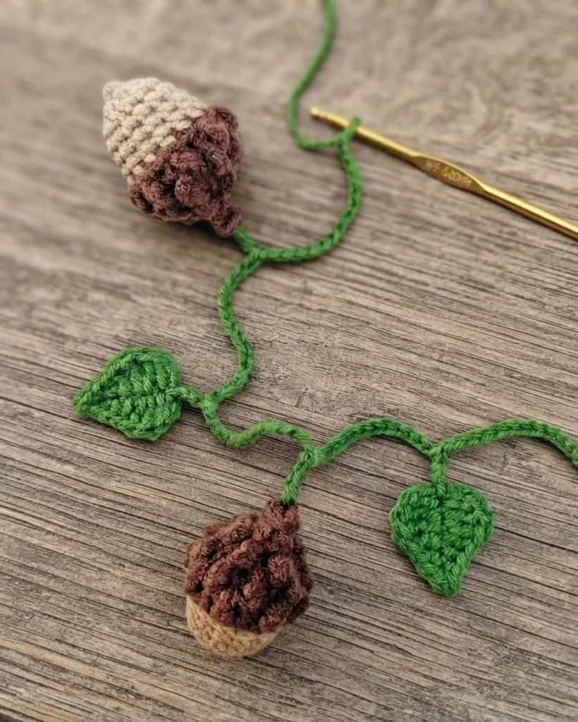 A crochet hook and green chain beside a partly assembled garland with crocheted acorns and leaves.