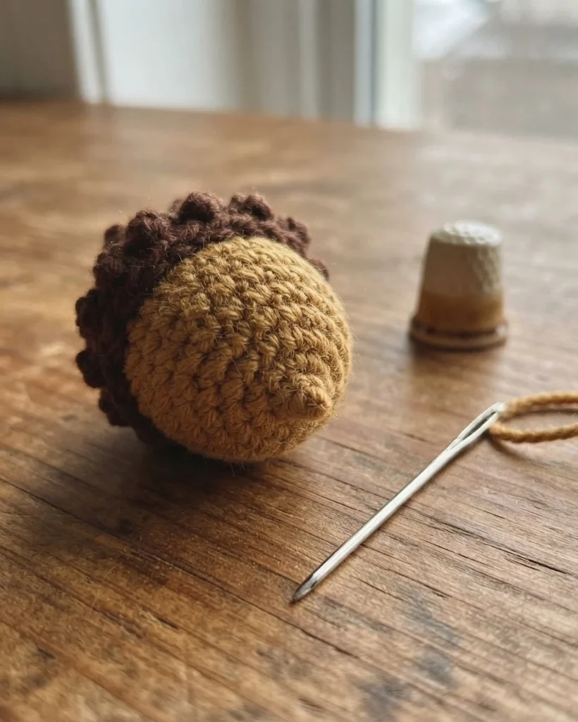 A small tan-and-brown crocheted acorn beside a yarn needle on a wooden table.