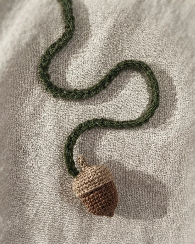 Marque-page crocheté en forme de gland brun et beige, relié à un long cordon vert foncé, posé sur du tissu beige.