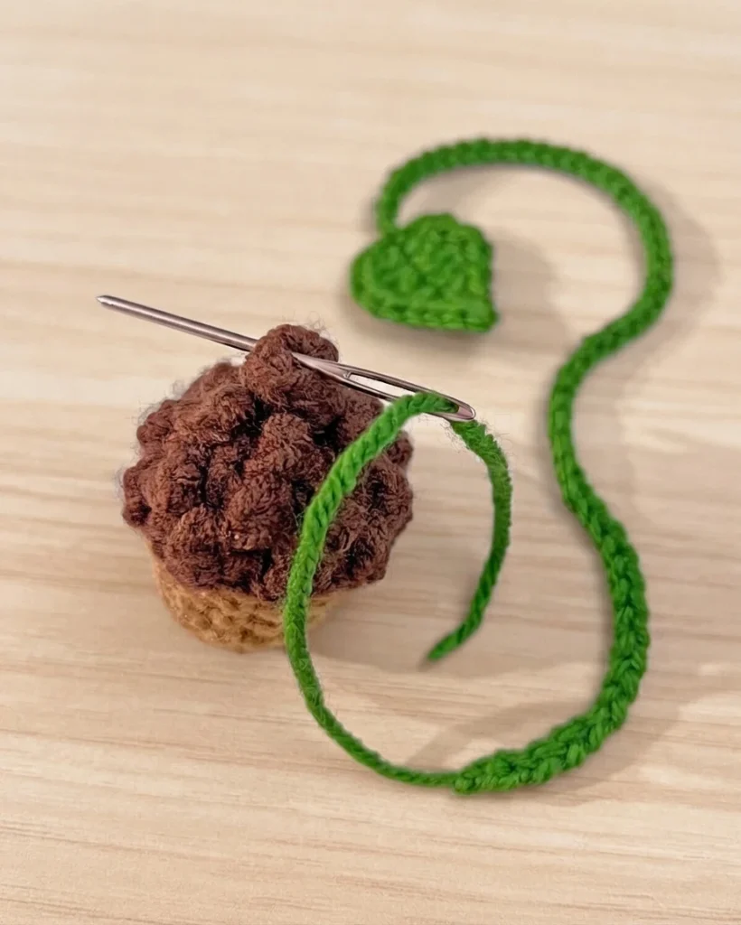 Une aiguille passe dans un petit gland au crochet brun et beige, près d’un cordon vert avec une feuille crochetée.