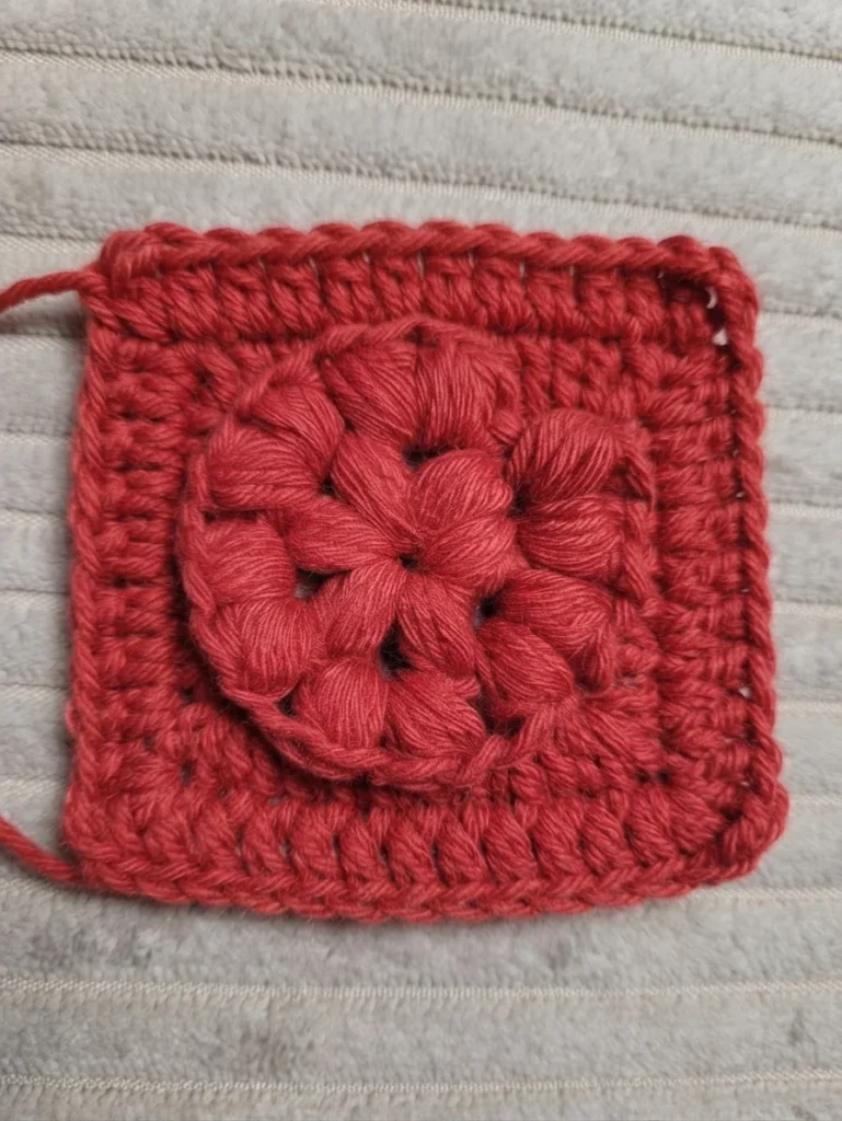 Red crochet swatch with a raised circular puff stitch motif in the center on light ribbed fabric