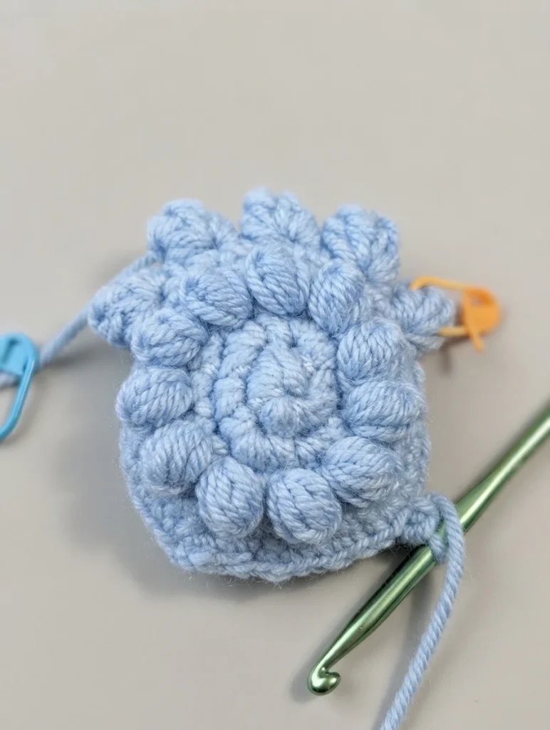 Light blue crochet piece with raised popcorn stitches, a green crochet hook and blue and orange stitch markers on a light surface