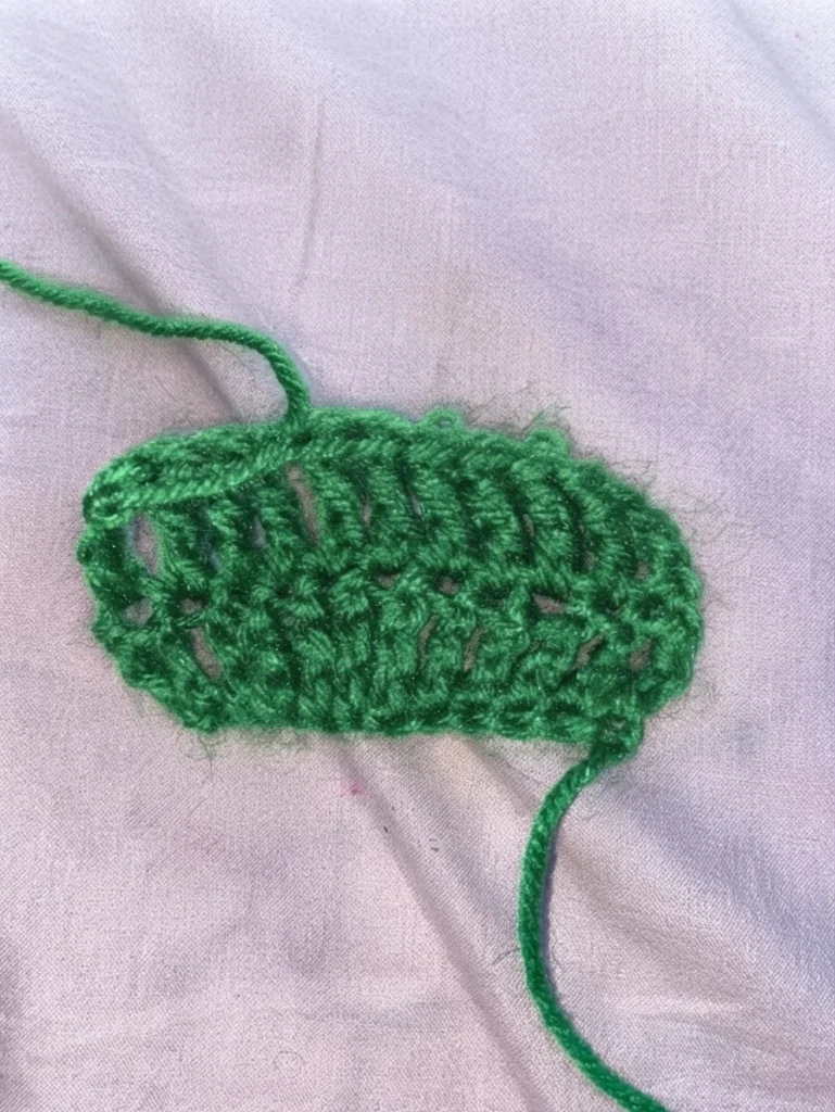 Green crochet swatch with tall open treble crochet stitches on light fabric, with loose yarn tails.