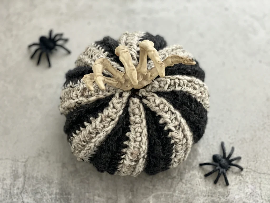 Black-and-cream striped crocheted pumpkin topped with a decorative skeleton hand, with two black spiders on a gray surface.