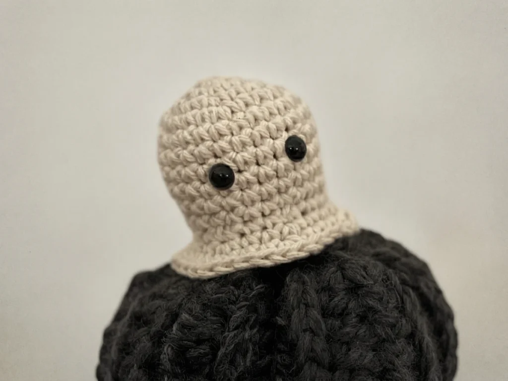 A small cream crocheted ghost with black eyes sits on a black crocheted pumpkin.