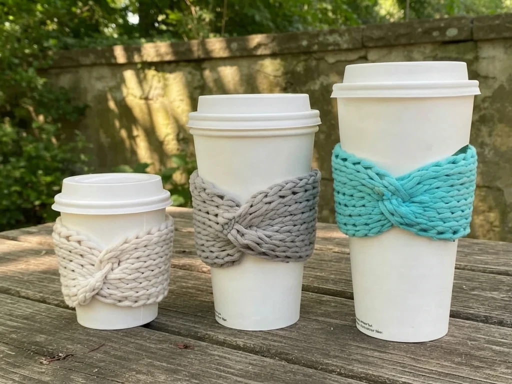 Three white takeaway cups with cream, gray, and turquoise crochet sleeves featuring crossed bow-like fronts, arranged outdoors on a wooden table.