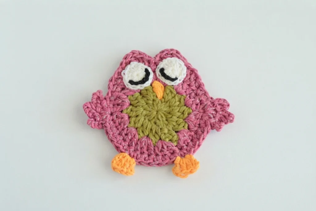 Single pink crochet owl coaster with white eyes, closed black stitched eyelids, a small orange beak, olive green center and orange feet, laid flat on a plain white surface.