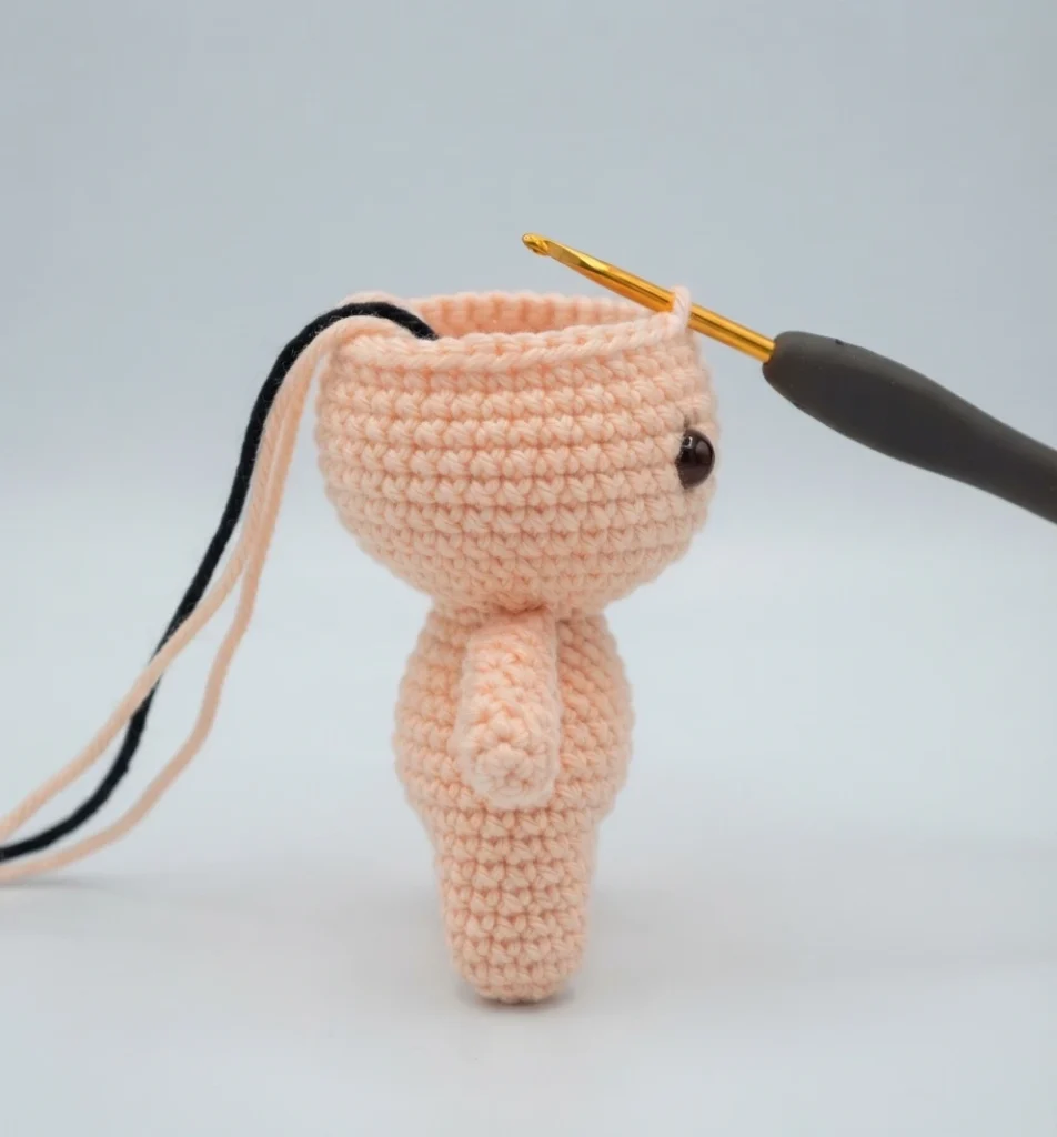 Small peach crocheted amigurumi piece with joined legs and a rounded open body, a gold crochet hook with a dark handle entering the top opening, long black and peach yarn tails trailing to the left, a dark safety eye visible on the right si