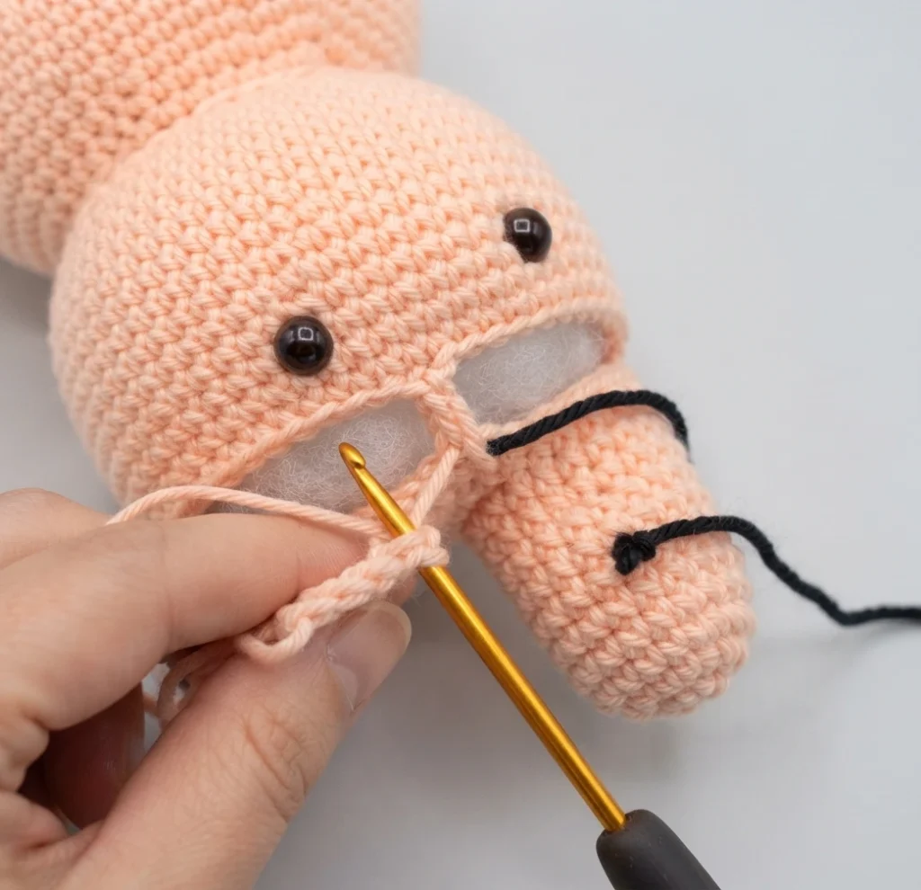 Hands crocheting a peach colored amigurumi piece with two black safety eyes and a stuffed muzzle, a gold crochet hook working the yarn with a black strand nearby.