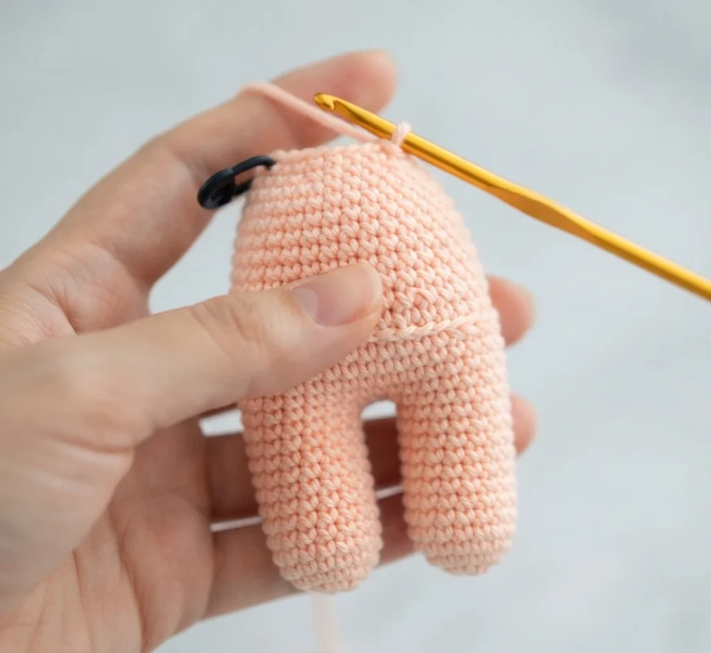 Hands holding a small peach-colored crocheted piece with two joined legs and a rounded body, a gold crochet hook inserted at the top edge and a black stitch marker clipped nearby, on a plain light background.
