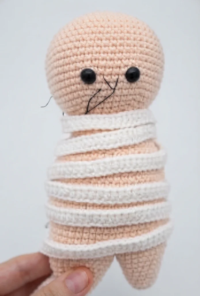 Crocheted amigurumi mummy doll with a pale skin tone body, two black safety eyes, embroidered mouth lines, and white crocheted strips wrapped diagonally around the torso and legs, held between fingers against a plain light background.