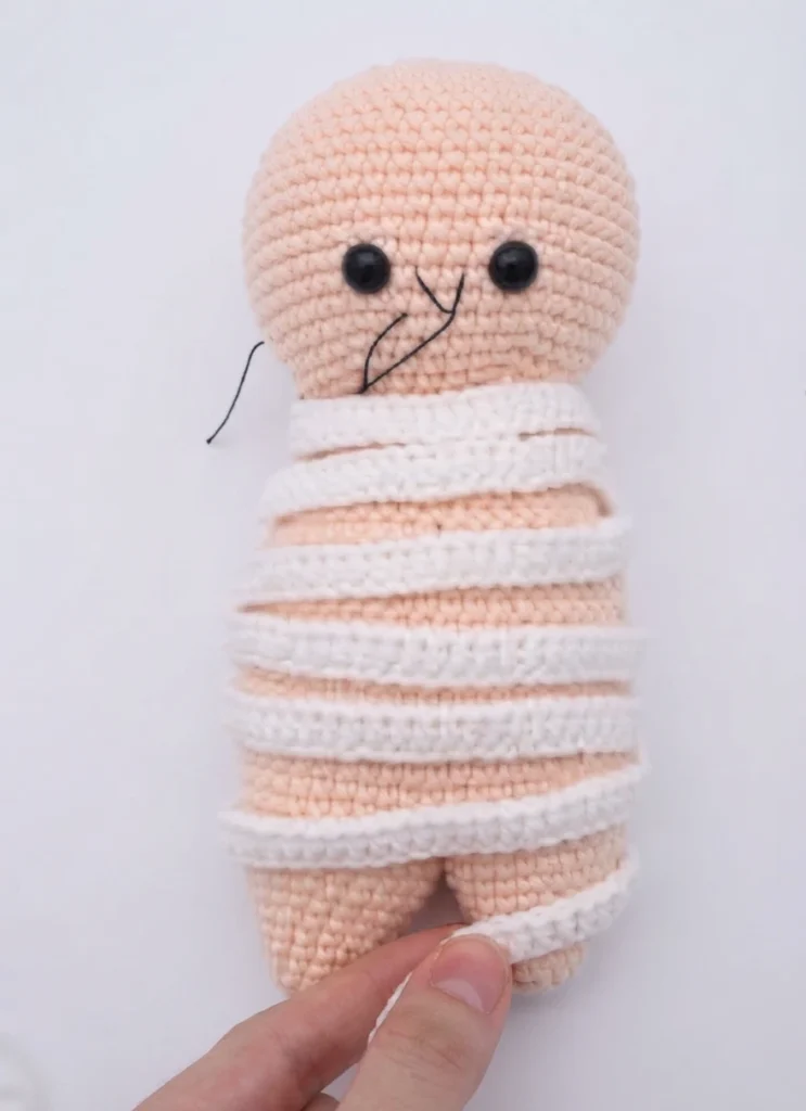 Crocheted mummy amigurumi in pale peach yarn with black bead eyes and stitched mouth, body wrapped in white crocheted strips, held by a hand against a plain white background.