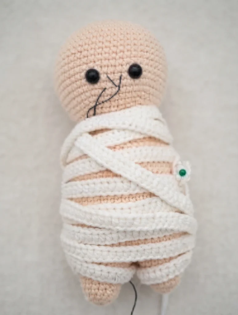 Crocheted mummy amigurumi doll with a peach skin tone, black safety eyes and a black stitched mouth, its body wrapped in cream crochet bands and secured with a green-headed pin, lying on a plain light background.
