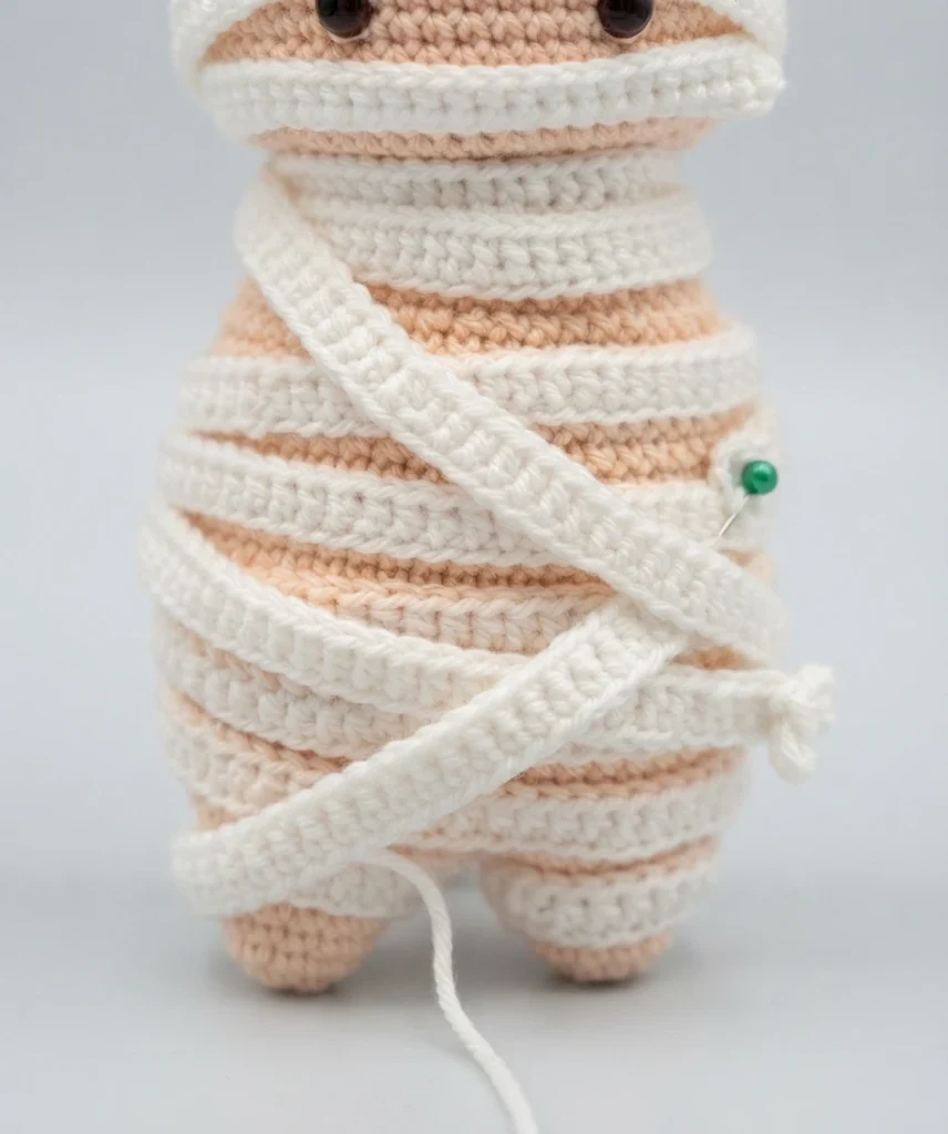 Crocheted mummy amigurumi body in skin tone yarn wrapped with narrow white crocheted strips in crisscross bands, held with a green sewing pin at the side, with a loose white yarn tail hanging below and two small feet showing at the bottom.