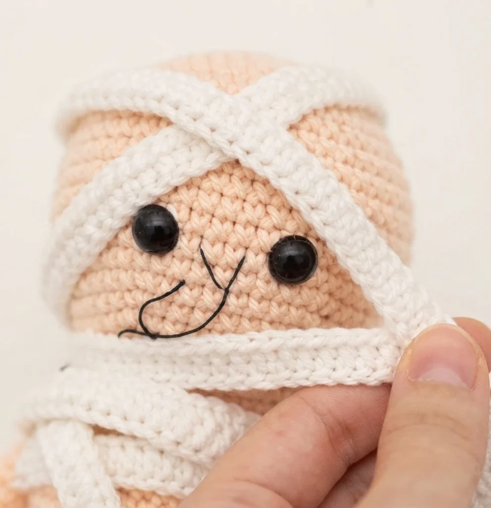 Close-up of a crocheted mummy doll head in peach yarn with black safety eyes and an embroidered smile, white crocheted wrapping bands crossing over the top and around the face opening, with a hand holding the loose end of the wrapping