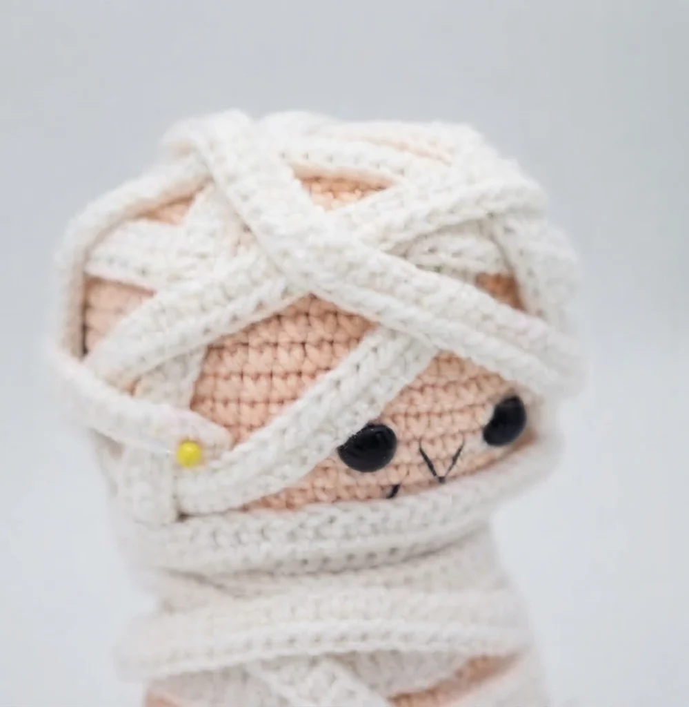 Close-up of a crocheted mummy doll head with white yarn strips criss-crossing over a beige face, two black safety eyes, a small stitched mouth and a yellow-headed sewing pin holding the wrap in place.