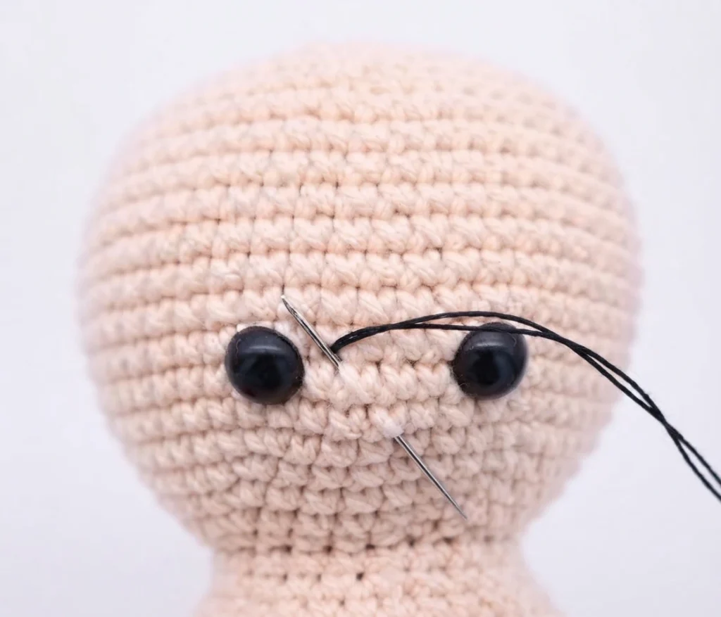 Close-up of a peach colored crocheted amigurumi head with two black safety eyes and black embroidery thread on a needle, showing the mouth being stitched between the eyes.