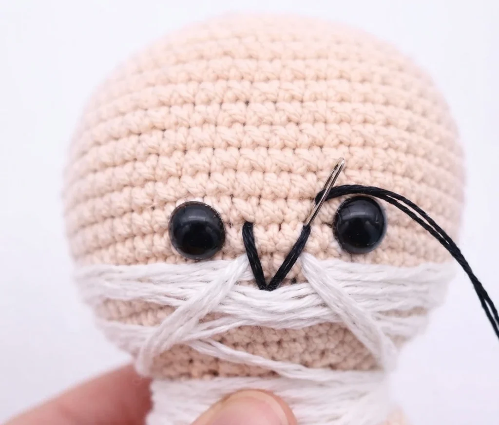 Crocheted doll head in light peach yarn with two black safety eyes, white yarn wrapped around the lower face and neck, and black embroidery thread forming a mouth with a needle and thread extending to the right.