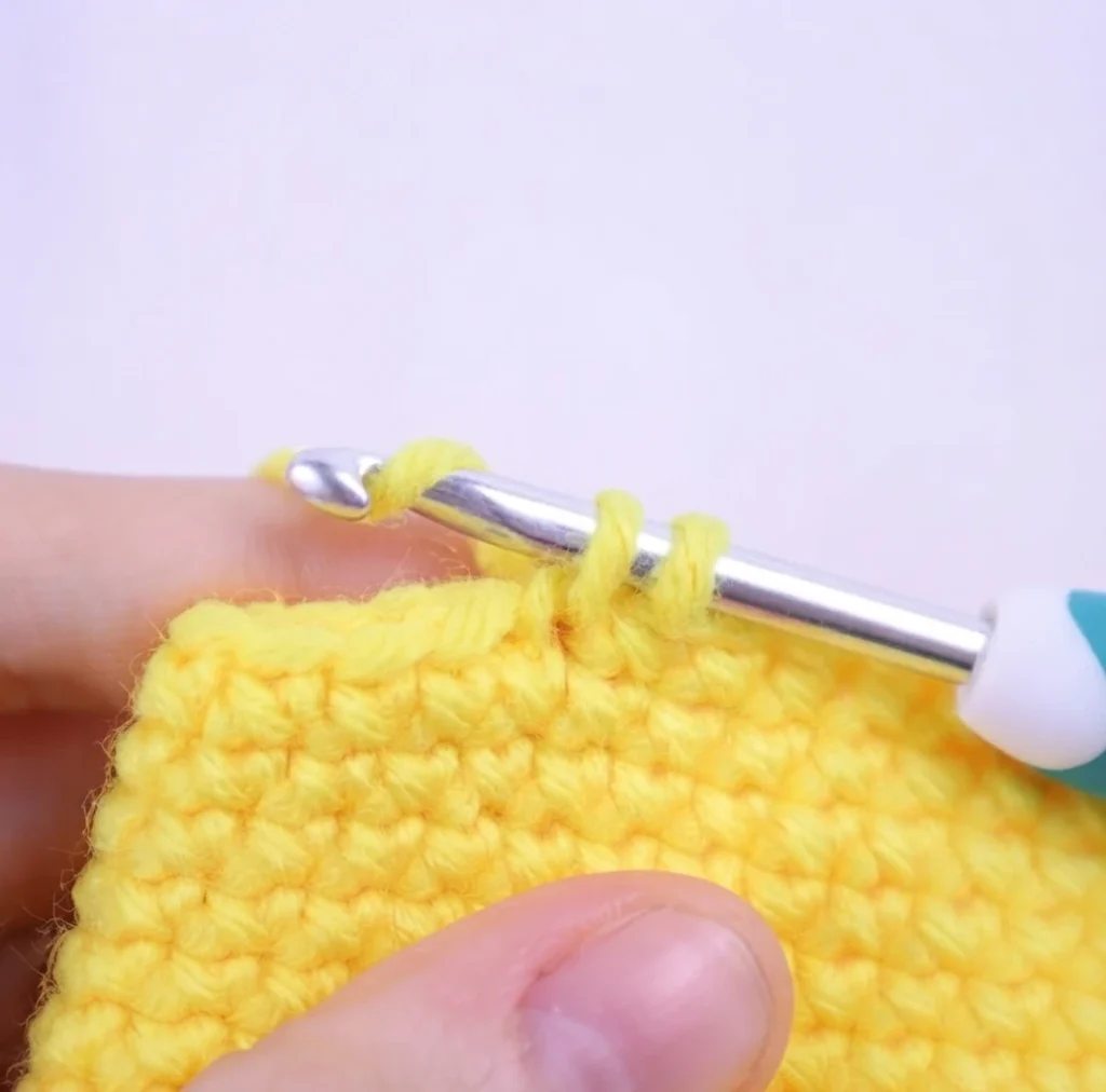 Close-up of a hand holding a yellow crocheted swatch while a metal crochet hook holds two yellow loops above the fabric, with a thumb resting on the lower edge against a pale lavender background.