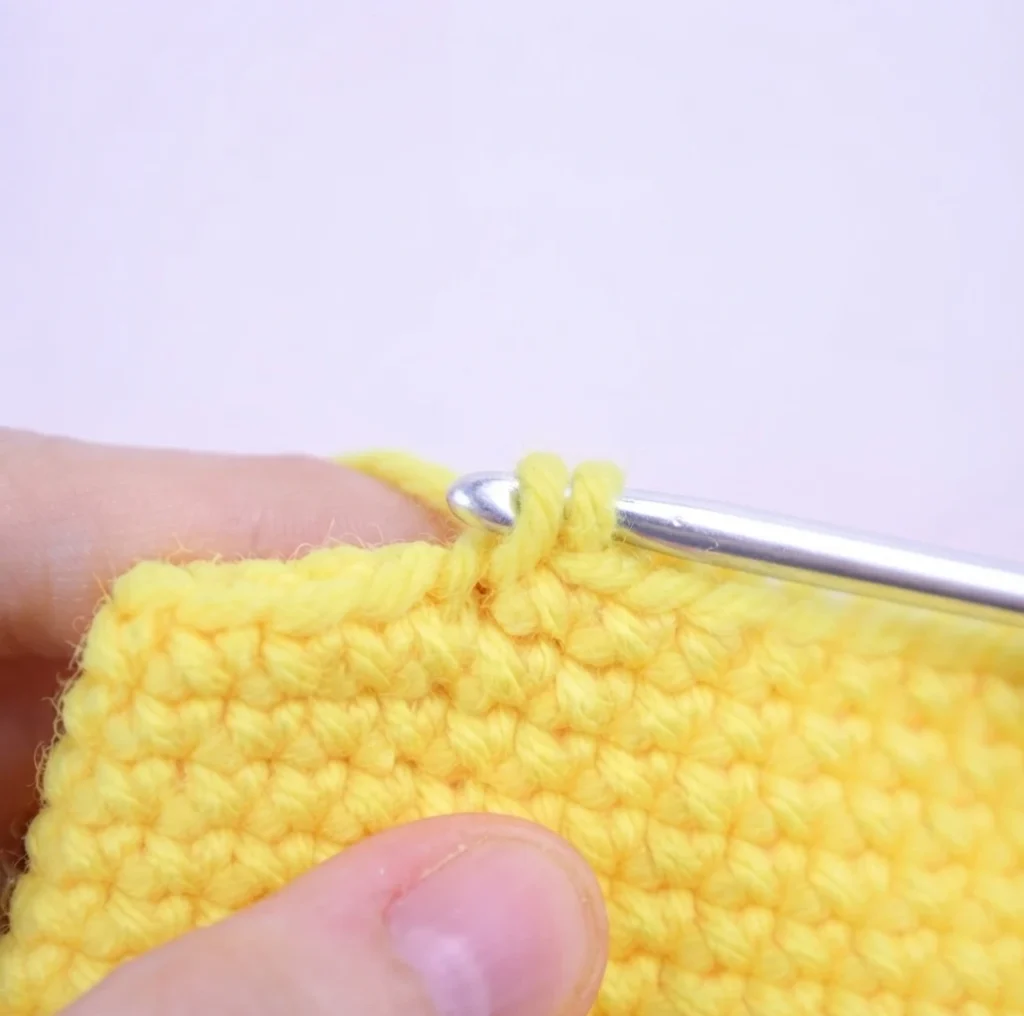 Close-up of hands holding a yellow crocheted swatch with a silver crochet hook inserted into the next stitch, working yarn held above the hook against a pale pink background.